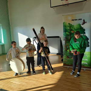 Grupka uczniów bierze udział w warsztatach dotyczących lasu. Dzieci trzymają w rękach różne przedmioty. Kliknięcie na zdjęcie spowoduje jego powiększenie do rozmiaru oryginalnego.