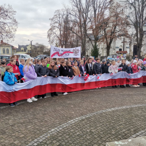 Uczniowie zgromadzeni w półkolu na rynku w Szczurowej z okazji obchodów 11 listopada.  Kliknięcie na zdjęcie spowoduje jego powiększenie do rozmiaru oryginalnego.