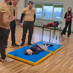 Strażacy uczą uczniów podstaw pierwszej pomocy, jedno z dzieci leży na materacu. Kliknięcie na zdjęcie spowoduje jego powiększenie do rozmiaru oryginalnego.