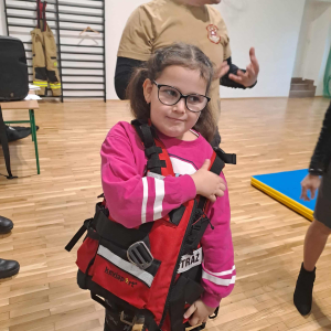 Uczennica klasy 1 mierzy kamizelkę ratunkową przywiezioną przez strażaków. Kliknięcie na zdjęcie spowoduje jego powiększenie do rozmiaru oryginalnego.