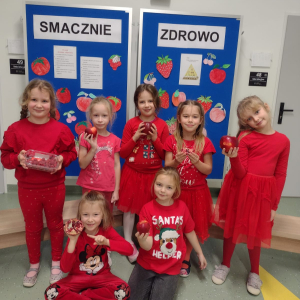 Dziewczynki z klasy 1 a prezentują czerwone owoce, w tle tablica z czerwonymi owocami. Kliknięcie na zdjęcie spowoduje jego powiększenie do rozmiaru oryginalnego.