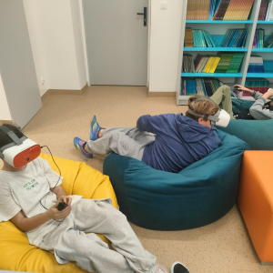 Uczniowie biorący udział w projekcie siedzą na pufach na głowach mają okulary VR. Kliknięcie na zdjęcie spowoduje jego powiększenie do rozmiaru oryginalnego.