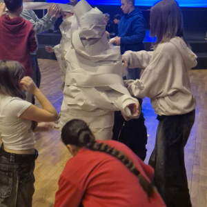 Młodzież owija papierem jedno z dzieci w czasie obchodów klasowych mikołajek. Klasa 7 b bawi się w krzesła - uczniowie chodzą wokół krzeseł. Kliknięcie na zdjęcie spowoduje jego  powiększenie do rozmiaru oryginalnego.