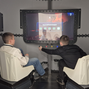 Dwóch uczniów siedzi na fotelach przed nimi ekran. Jedne z nich naciska przyciski. Kliknięcie na zdjęcie spowoduje jego powiększenie do rozmiaru oryginalnego.