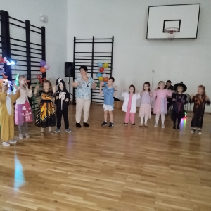 Dzieci wraz z nauczycielami stoją w półkole, bawią się na balu karnawałowym. Kliknięcie na zdjęcie spowoduje jego powiększenie do rozmiaru oryginalnego.