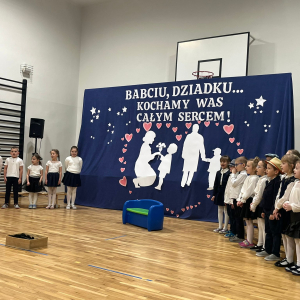 Przedszkolaki stoją w półkolu w trakcie występu dla dziadków. Kliknięcie na zdjęcie spowoduje jego powiększenie do rozmiaru oryginalnego.