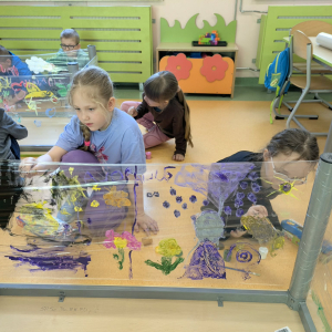 Przedszkolaki malują farbkami na foliach przymocowanych do odwróconych stolików. Kliknięcie na zdjęcie spowoduje jego powiększenie do rozmiaru oryginalnego.