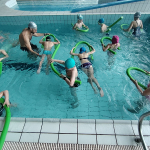 Uczniowie biorący udział w projekcie pływają pod okiem instruktora w basenie. Dzieci trzymają w rękach gumiane węże do nauki pływania. Kliknięci na zdjęcie spowoduje jego powiększenie do rozmiaru oryginalnego.
