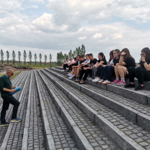 Młodzież z klas 8 siedzi na kamiennych schodach na terenie Nazistowskiego Obozu Zagłady w Oświęcimiu. Słucha wykładu przewodnika. Kliknięcie na zdjęcie spowoduje jego powiększenie do rozmiaru oryginalnego.