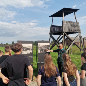 Młodzież wraz z przewodnikiem zwiedza teren Nazistowskiego Obozu Zagłady w Oświęcimiu. Uczestnicy wycieczki stoją przy jednej ze wież strażniczych. Kliknięcie na zdjęcie spowoduje jego powiększenie do oryginalnego rozmiaru.