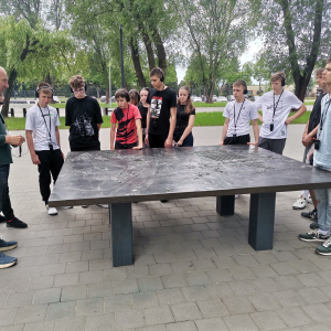 Przewodnik wraz z uczniami stoi przy kamiennym stole znajdującym się na terenie Nazistowskiego Obozu Zagłady w Oświęcimiu. Kliknięcie na zdjęcie spowoduje jego powiększenie do rozmiaru oryginalnego.