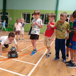 Dzieci z klasy 1 a obserwują jeżdżącego robota w ramach warsztatów z robotyki. Kliknięcie na zdjęcie spowoduje jego powiększenie do rozmiaru oryginalnego.