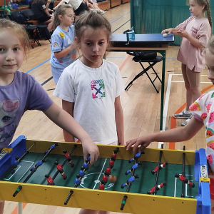 Grupka dziewczynek z klasy 1 gra w piłkarzyki na hali sportowej. Dzieci bawią się na Dniu Dziecka. Kliknięcie na zdjęcie spowoduje jego powiększenie do rozmiaru oryginalnego.