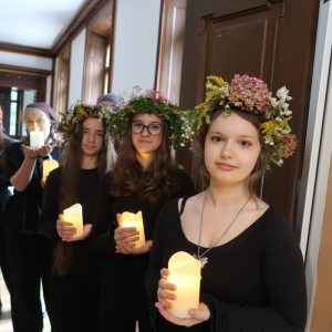 Uczennice klas 8 stoją w szeregu ubrane w wianki, trzymają świece w rękach. Kliknięcie na zdjęcie spowoduje jego powiększenie do rozmiaru oryginalnego.